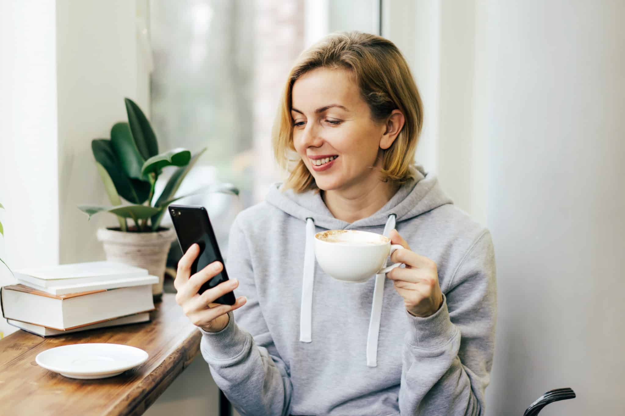 A woman drinks coffee and communicates on a messenger on her phone in a bright cafe by the window.